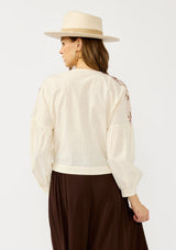 [Color: Vanilla/Rust] A back facing image of a blonde model wearing an off white embroidered wrap top with dropped shoulders, long sleeves, and a side tie closure. An effortless boho top for the fall season. 