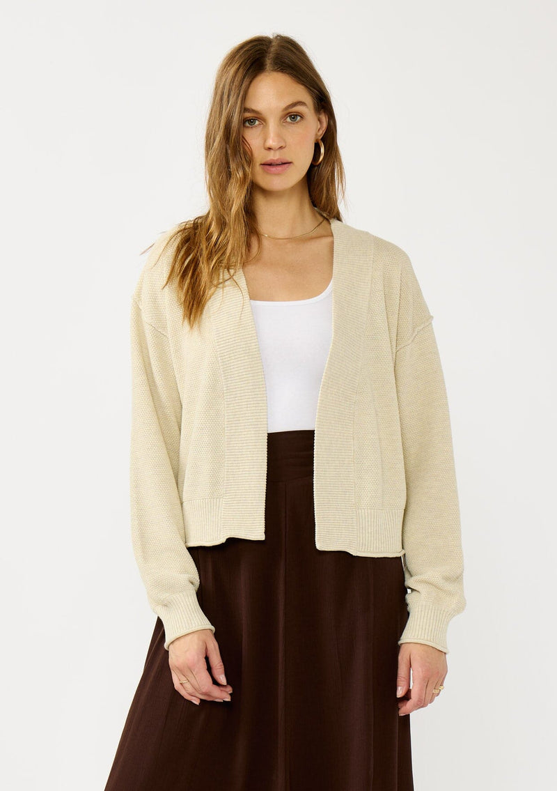 [Color: Heather Natural] A front facing image of a blonde model wearing a beige casual, lightweight cardigan with an open front, ribbed trim, and rolled edges. A classic staple cardigan for transitional styling. 