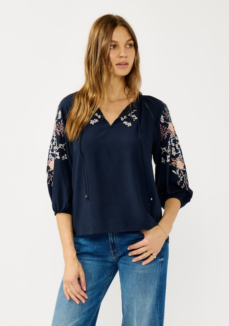 [Color: Eclipse/Coral] A front facing image of a blonde model wearing a dark blue embroidered blouse with 3/4 length sleeves,  a split v-neckline with tie details, and lattice trim accents. 
