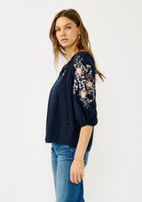 [Color: Eclipse/Coral] A side facing image of a blonde model wearing a dark blue embroidered blouse with 3/4 length sleeves,  a split v-neckline with tie details, and lattice trim accents. 