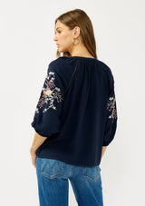 [Color: Eclipse/Coral] A back facing image of a blonde model wearing a dark blue embroidered blouse with 3/4 length sleeves,  a split v-neckline with tie details, and lattice trim accents. 