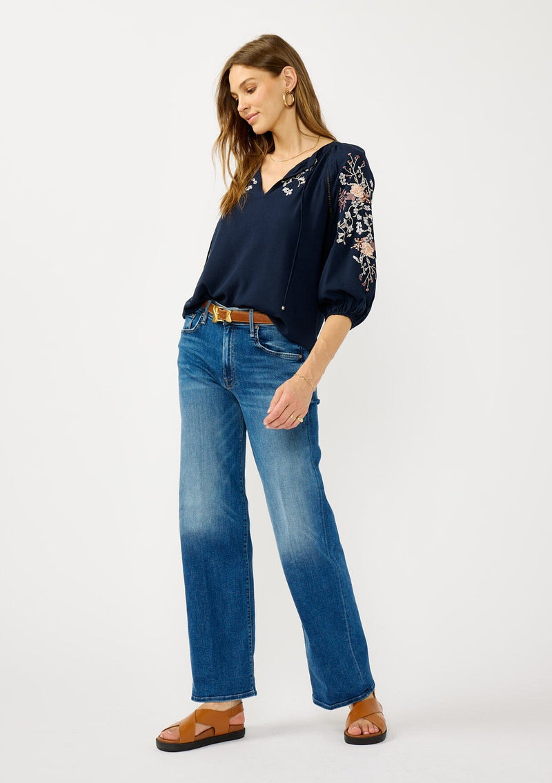 [Color: Eclipse/Coral] A front facing image of a blonde model wearing a dark blue embroidered blouse with 3/4 length sleeves,  a split v-neckline with tie details, and lattice trim accents. 