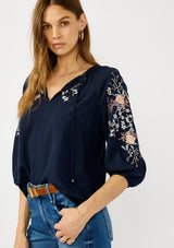 [Color: Eclipse/Coral] A front facing image of a blonde model wearing a dark blue embroidered blouse with 3/4 length sleeves,  a split v-neckline with tie details, and lattice trim accents. 