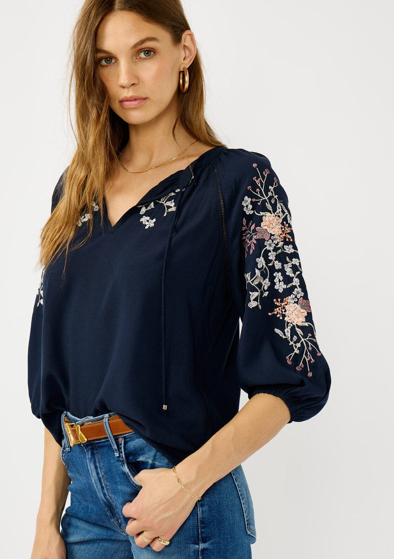 [Color: Eclipse/Coral] A front facing image of a blonde model wearing a dark blue embroidered blouse with 3/4 length sleeves,  a split v-neckline with tie details, and lattice trim accents. 