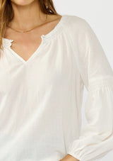 [Color: White] A detail image of a blonde model wearing a white crinkled cotton blouse with long bishop sleeves, shirred detail along the sleeves, a split v-neckline with button closure. A year round staple for any season. 