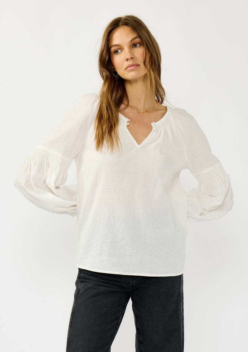 [Color: White] A front facing image of a blonde model wearing a white crinkled cotton blouse with long bishop sleeves, shirred detail along the sleeves, a split v-neckline with button closure. A year round staple for any season. 