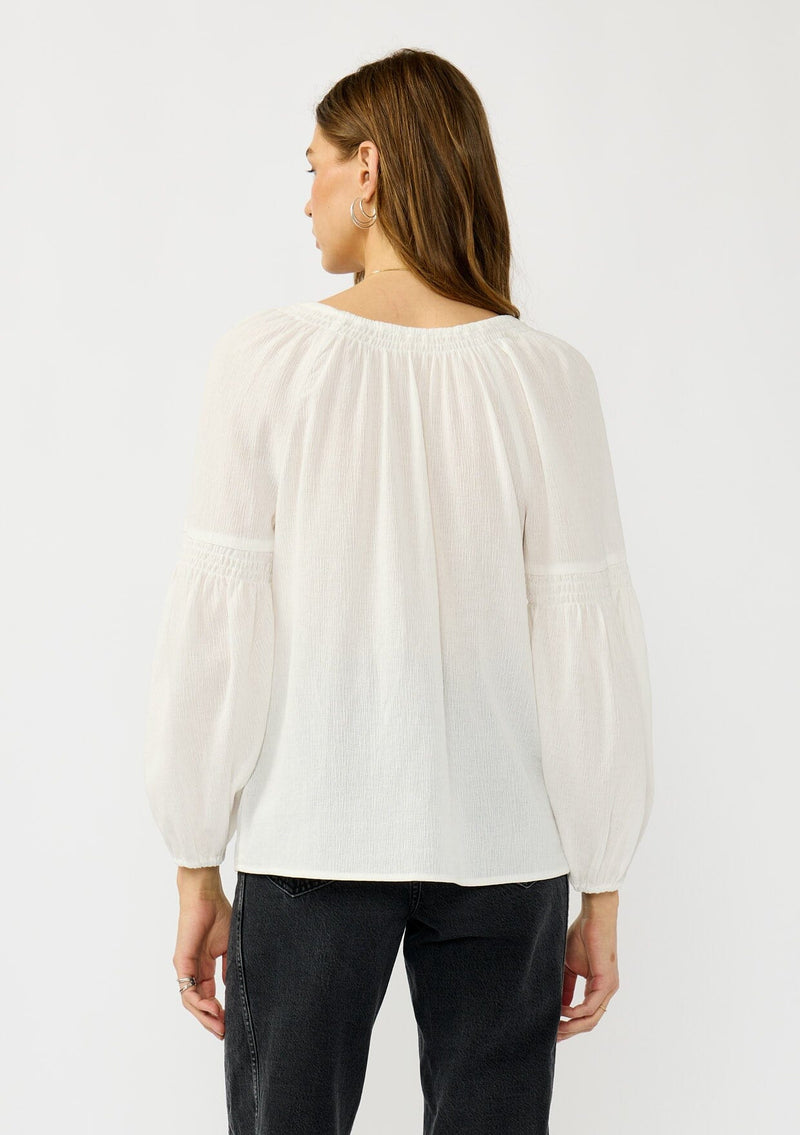 [Color: White] A back facing image of a blonde model wearing a white crinkled cotton blouse with long bishop sleeves, shirred detail along the sleeves, a split v-neckline with button closure. A year round staple for any season. 