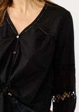 [Color: Black] A detail image of a blonde model wearing a black lightweight cotton-blend top with a tie front detail, button front, and 3/4 length sleeves with embroidered and cutout details. 