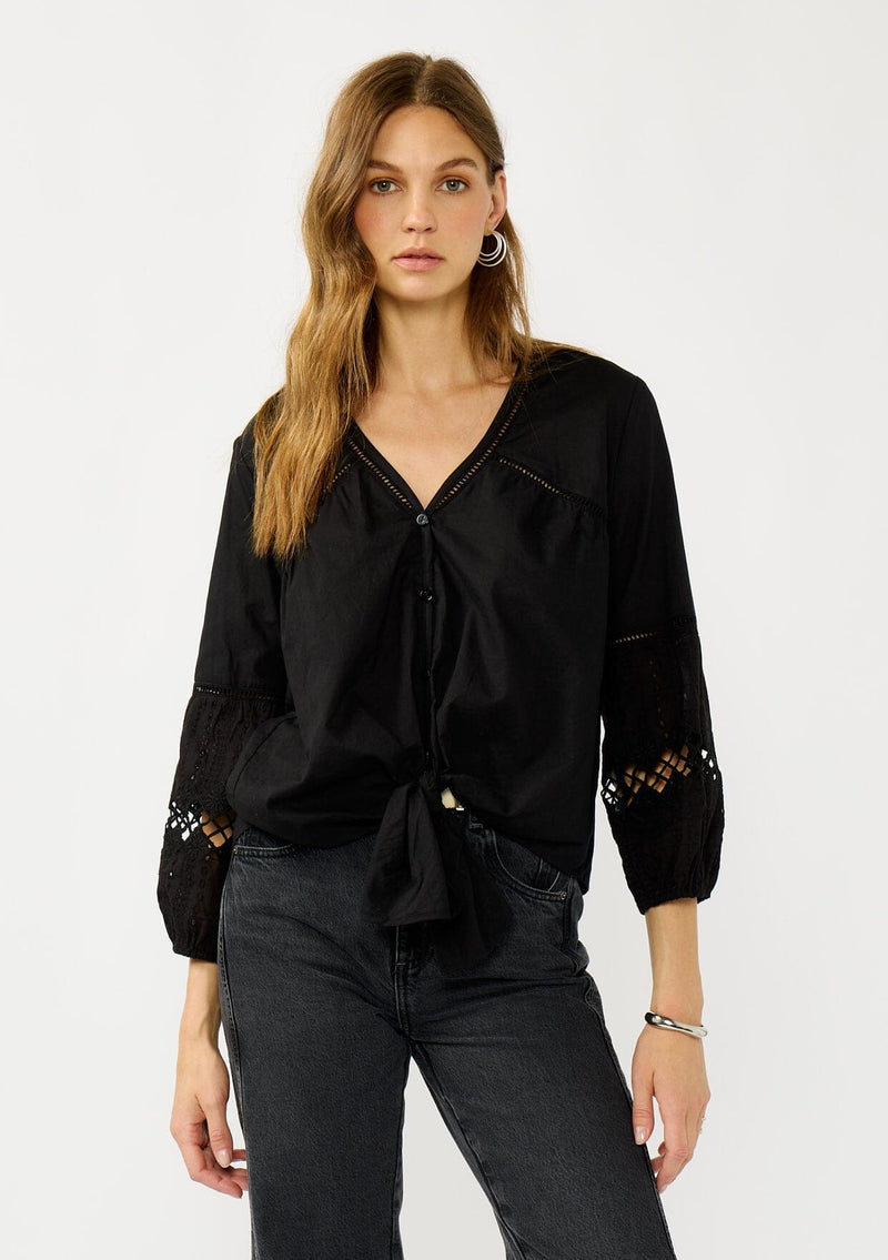 [Color: Black] A front facing image of a blonde model wearing a black lightweight cotton-blend top with a tie front detail, button front, and 3/4 length sleeves with embroidered and cutout details. 