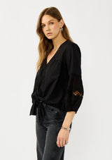 [Color: Black] A side facing image of a blonde model wearing a black lightweight cotton-blend top with a tie front detail, button front, and 3/4 length sleeves with embroidered and cutout details. 