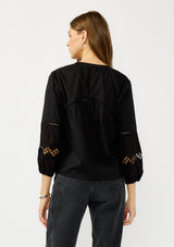 [Color: Black] A back facing image of a blonde model wearing a black lightweight cotton-blend top with a tie front detail, button front, and 3/4 length sleeves with embroidered and cutout details. 