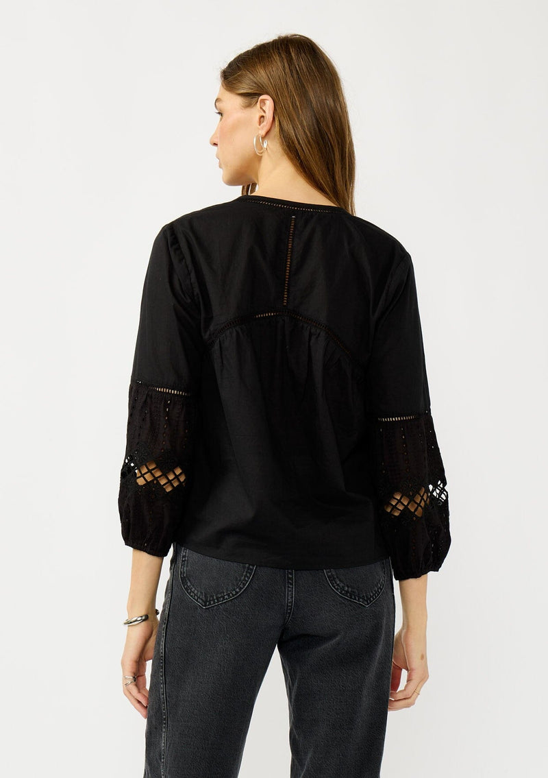 [Color: Black] A back facing image of a blonde model wearing a black lightweight cotton-blend top with a tie front detail, button front, and 3/4 length sleeves with embroidered and cutout details. 