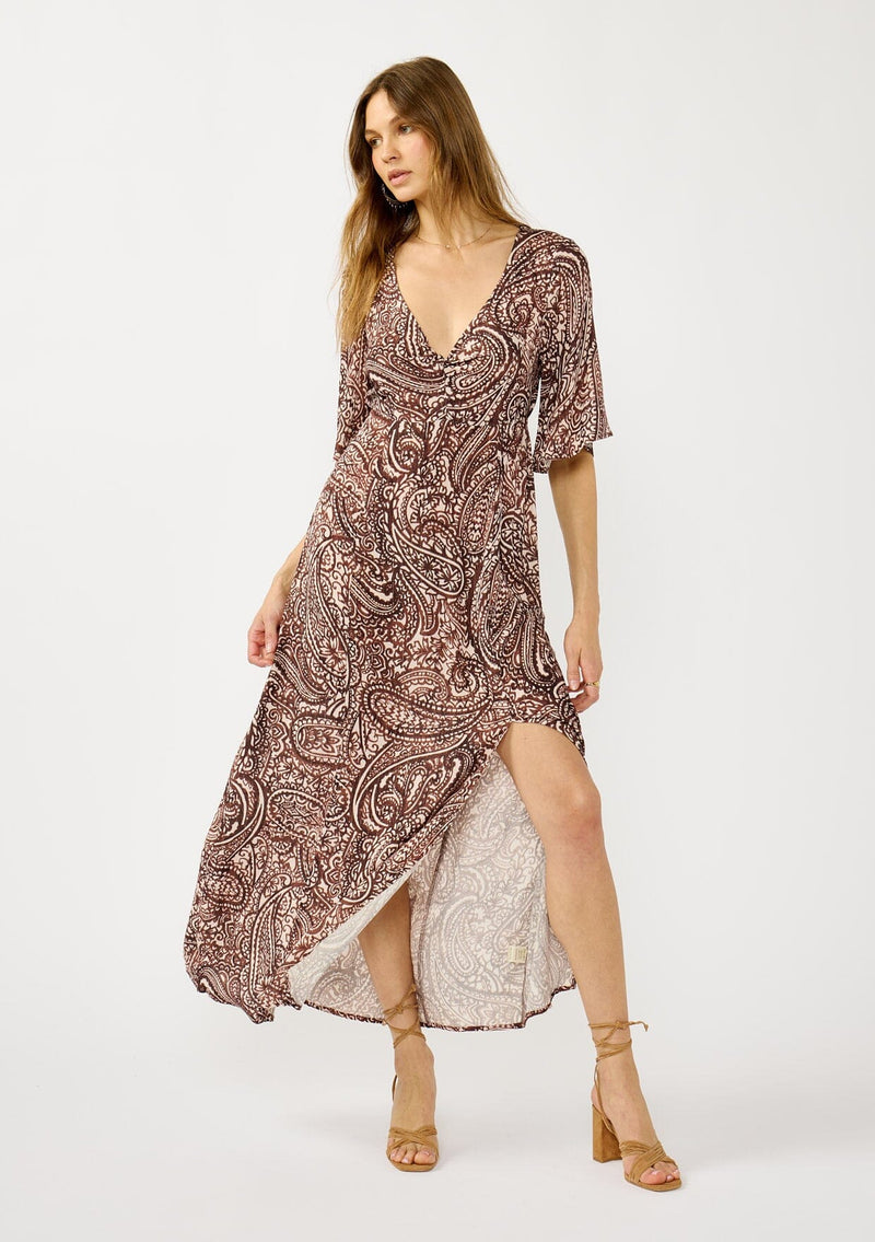 [Color: Natural/Brown] A front facing image of a blonde model wearing a brown paisley maxi dress with a deep v-neckline, flowy short sleeves, twist front, and a side slit. A fall bohemian maxi dress for special occasions. 