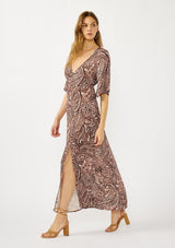 [Color: Natural/Brown] A side facing image of a blonde model wearing a brown paisley maxi dress with a deep v-neckline, flowy short sleeves, twist front, and a side slit. A fall bohemian maxi dress for special occasions. 