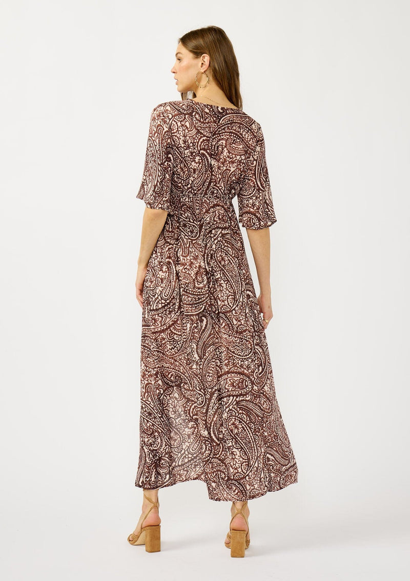 [Color: Natural/Brown] A back facing image of a blonde model wearing a brown paisley maxi dress with a deep v-neckline, flowy short sleeves, twist front, and a side slit. A fall bohemian maxi dress for special occasions. 