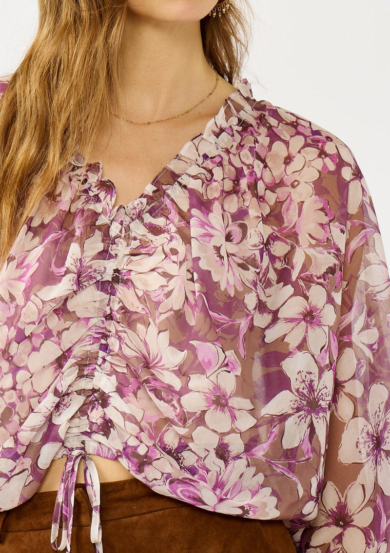 [Color: Mocha/Lilac] A detail image of a blonde model wearing a billowy purple floral blouse with voluminous sleeves, a ruched front, ruffled v-neckline, and an elastic waist. A flowy chiffon top for the fall season. 