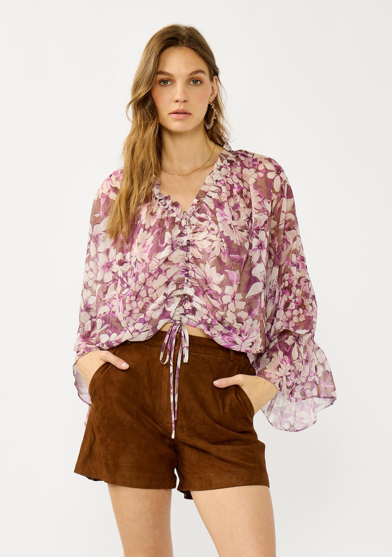 [Color: Mocha/Lilac] A front facing image of a blonde model wearing a billowy purple floral blouse with voluminous sleeves, a ruched front, ruffled v-neckline, and an elastic waist. A flowy chiffon top for the fall season. 