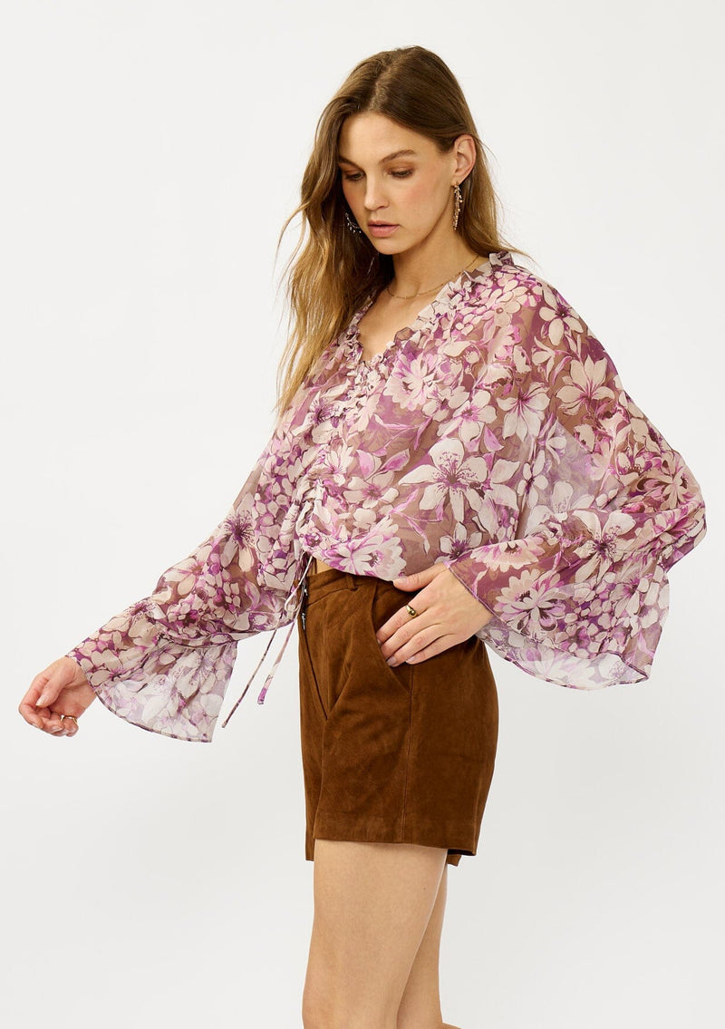 [Color: Mocha/Lilac] A side facing image of a blonde model wearing a billowy purple floral blouse with voluminous sleeves, a ruched front, ruffled v-neckline, and an elastic waist. A flowy chiffon top for the fall season. 