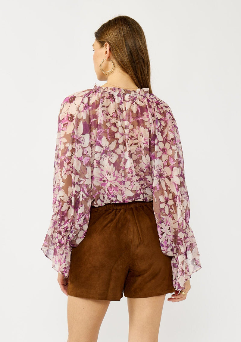 [Color: Mocha/Lilac] A back facing image of a blonde model wearing a billowy purple floral blouse with voluminous sleeves, a ruched front, ruffled v-neckline, and an elastic waist. A flowy chiffon top for the fall season. 