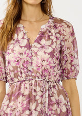 [Color: Mocha/Lilac] A detail image of a blonde model wearing a purple floral chiffon mini dress with puff sleeves, a split v-neckline, button front detail, and an elastic waist with belt tie. A flowy fall mini dress for special occasions. 