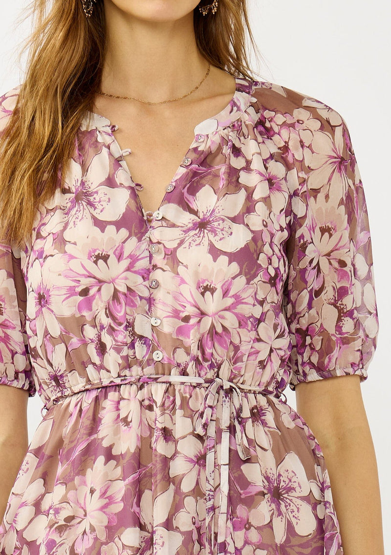 [Color: Mocha/Lilac] A detail image of a blonde model wearing a purple floral chiffon mini dress with puff sleeves, a split v-neckline, button front detail, and an elastic waist with belt tie. A flowy fall mini dress for special occasions. 