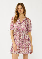 [Color: Mocha/Lilac] A front facing image of a blonde model wearing a purple floral chiffon mini dress with puff sleeves, a split v-neckline, button front detail, and an elastic waist with belt tie. A flowy fall mini dress for special occasions. 