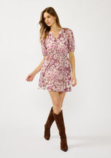 [Color: Mocha/Lilac] A front facing image of a blonde model wearing a purple floral chiffon mini dress with puff sleeves, a split v-neckline, button front detail, and an elastic waist with belt tie. A flowy fall mini dress for special occasions. 