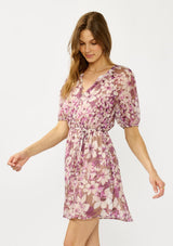 [Color: Mocha/Lilac] A side facing image of a blonde model wearing a purple floral chiffon mini dress with puff sleeves, a split v-neckline, button front detail, and an elastic waist with belt tie. A flowy fall mini dress for special occasions. 