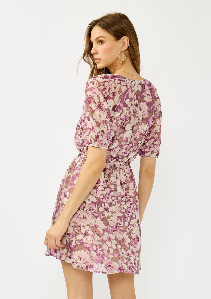 [Color: Mocha/Lilac] A back facing image of a blonde model wearing a purple floral chiffon mini dress with puff sleeves, a split v-neckline, button front detail, and an elastic waist with belt tie. A flowy fall mini dress for special occasions. 