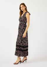 [Color: Black/Pink] A side facing image of a blonde model wearing a black and pink floral print maxi dress with flutter cap sleeves, a v-neckline, button front, smocked back, and sheer tiered layers. A special occasion dress for the fall season. 