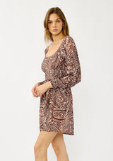 [Color: Natural/Brown] A side facing image of a blonde model wearing a brown paisley mini dress with gold thread details, long sleeves, smocked cuffs, smocked waistline, and a comfortable scooped neckline. A fall bohemian mini dress for casual occasions. 