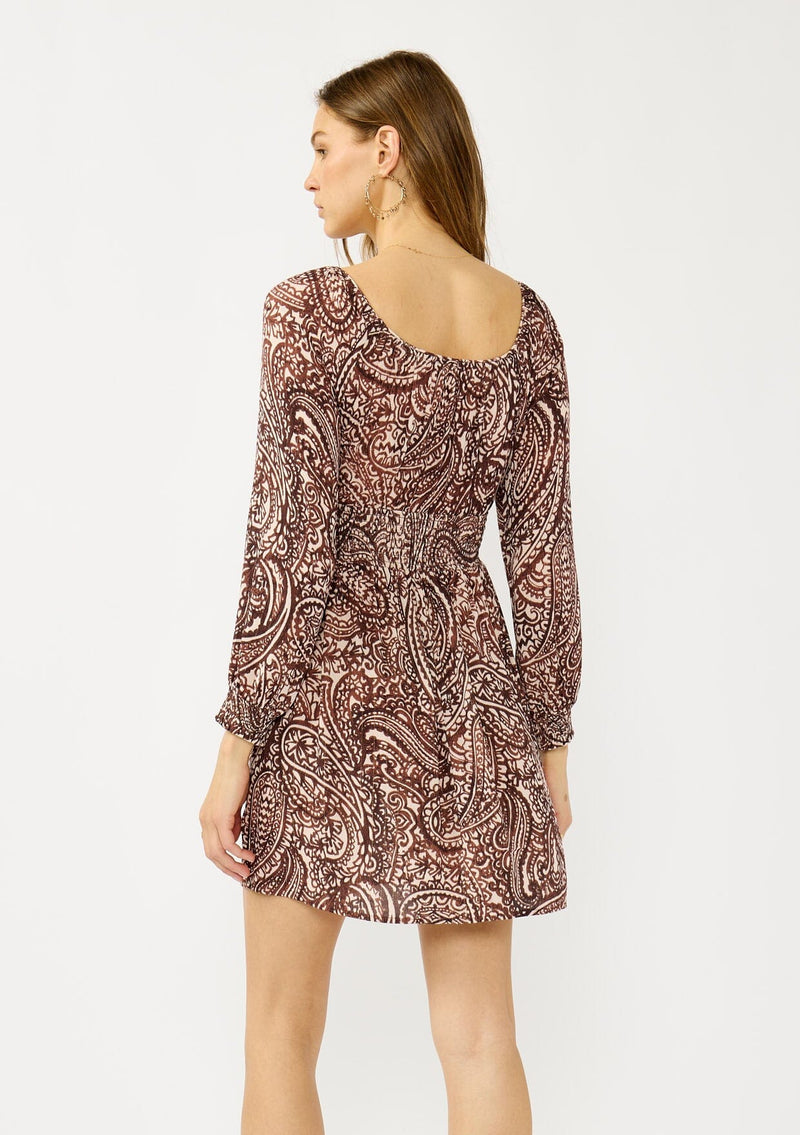 [Color: Natural/Brown] A back facing image of a blonde model wearing a brown paisley mini dress with gold thread details, long sleeves, smocked cuffs, smocked waistline, and a comfortable scooped neckline. A fall bohemian mini dress for casual occasions. 