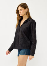[Color: Graphite] A side facing image of a blonde model wearing a black eyelet shirt with a collared neckline, button front, and button cuff closure.