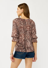 [Color: Natural/Brown] A back facing image of a blonde model wearing a brown paisley blouse with gold metallic threads, a split v-neckline, and 3/4 length sleeves with ruffled elastic cuffs. A relaxed boho top for the fall season. 