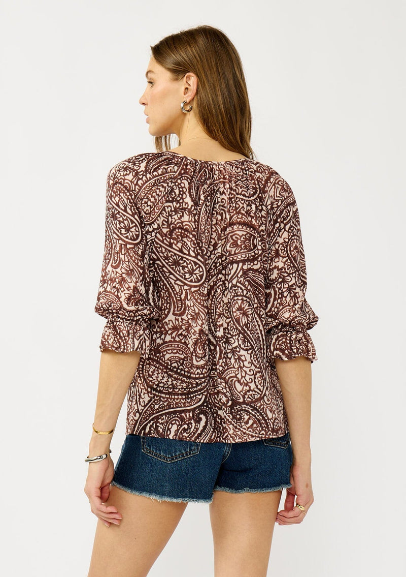 [Color: Natural/Brown] A back facing image of a blonde model wearing a brown paisley blouse with gold metallic threads, a split v-neckline, and 3/4 length sleeves with ruffled elastic cuffs. A relaxed boho top for the fall season. 