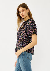 [Color: Black/Pink] A side facing image of a black and pink floral print top with puff sleeves, a split v-neckline with ties, shirred details, and lace trims. A day to night versatile top for the fall season. 