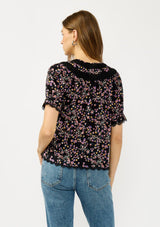 [Color: Black/Pink] A back facing image of a black and pink floral print top with puff sleeves, a split v-neckline with ties, shirred details, and lace trims. A day to night versatile top for the fall season. 