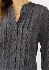 [Color: Navy/Taupe] A detail image of a blonde model wearing a dark navy checkered blouse with a round neckline, button front, and center front pintuck details. A casual fall piece with a polished touch for work and casual outings. 