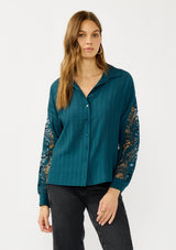 [Color: Teal] A front facing image of a blonde model wearing a striped jacquard teal shirt with lace cutouts, a button front, and collared neckline. A cool and polished blouse for the fall season. 