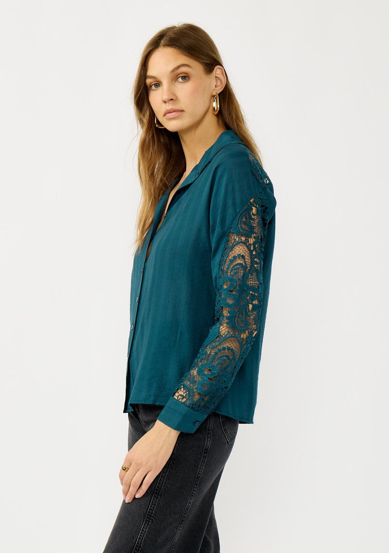 [Color: Teal] A side facing image of a blonde model wearing a striped jacquard teal shirt with lace cutouts, a button front, and collared neckline. A cool and polished blouse for the fall season. 