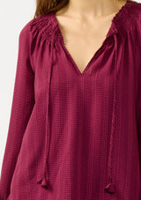 [Color: Wine] A detail image of a blonde model wearing a wine checkered blouse with a split smocked neckline, tie neck detail, a hook and eye  closure, and long sleeves with smocked cuffs. 