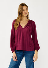 [Color: Wine] A front facing image of a blonde model wearing a wine checkered blouse with a split smocked neckline, tie neck detail, a hook and eye  closure, and long sleeves with smocked cuffs. 