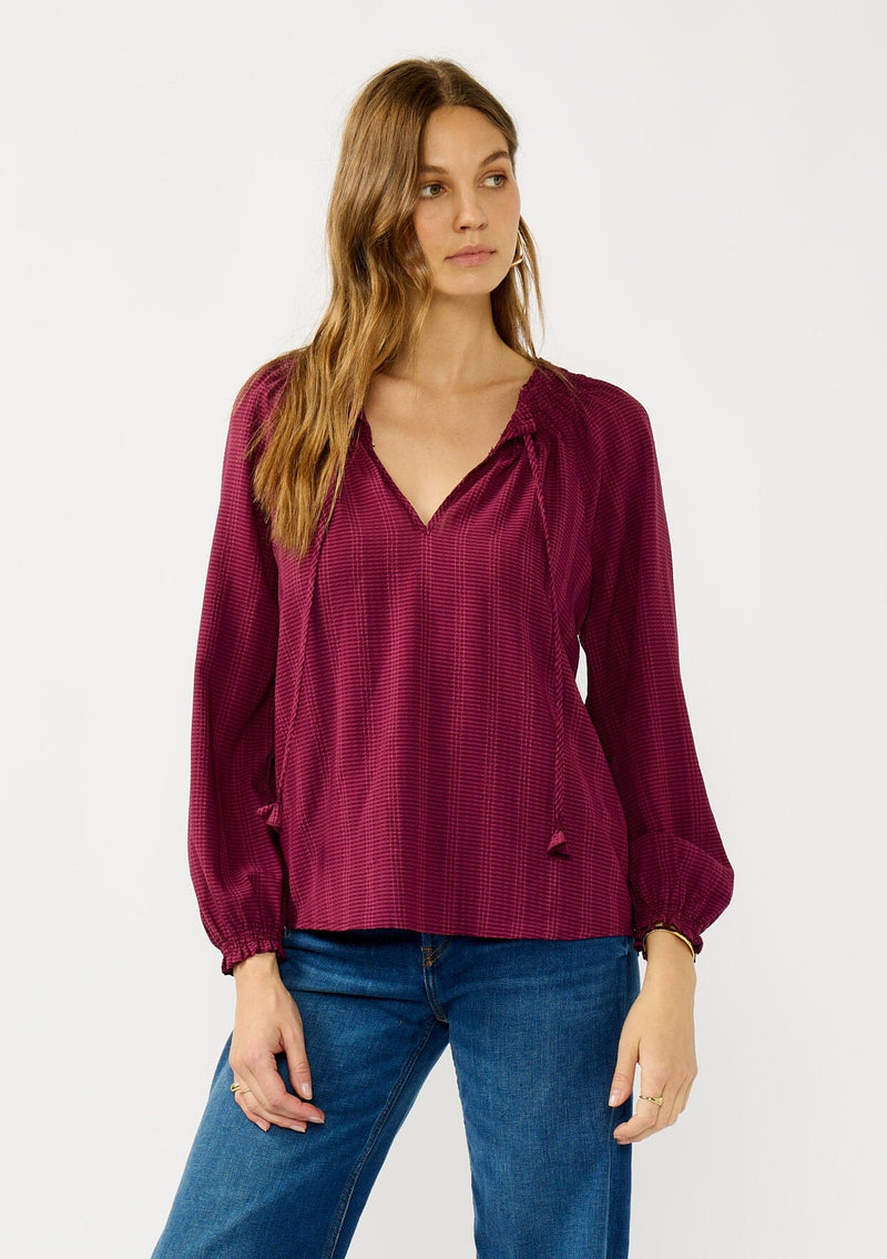 [Color: Wine] A front facing image of a blonde model wearing a wine checkered blouse with a split smocked neckline, tie neck detail, a hook and eye  closure, and long sleeves with smocked cuffs. 