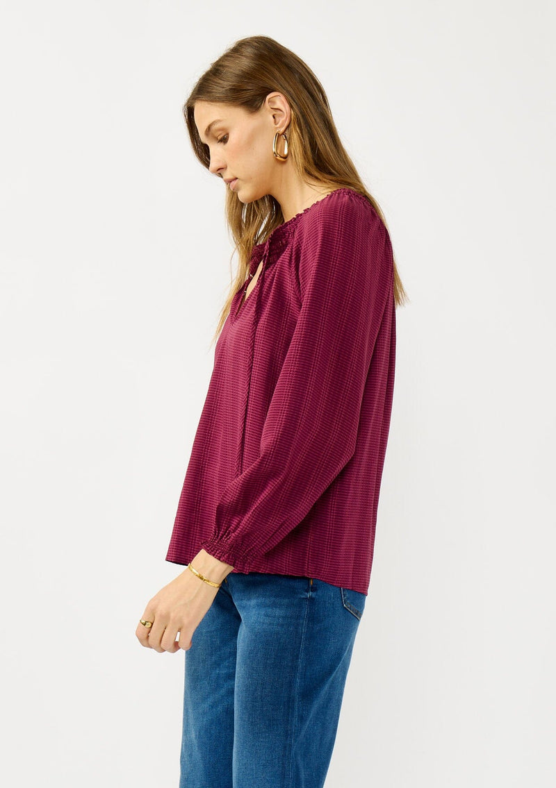 [Color: Wine] A side facing image of a blonde model wearing a wine checkered blouse with a split smocked neckline, tie neck detail, a hook and eye  closure, and long sleeves with smocked cuffs. 