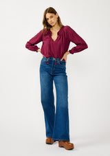 [Color: Wine] A front facing image of a blonde model wearing a wine checkered blouse with a split smocked neckline, tie neck detail, a hook and eye  closure, and long sleeves with smocked cuffs. 