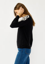 [Color: Black] A side facing image of a blonde model wearing a black pullover sweater with pearl embellishment on the shoulder. A classic and feminine sweater for the fall season. 