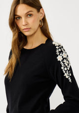 [Color: Black] A front facing image of a blonde model wearing a black pullover sweater with pearl embellishment on the shoulder. A classic and feminine sweater for the fall season. 