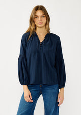 [Color: Midnight] A front facing image of a blonde model wearing a blue long sleeve peasant blouse with a subtle checkered pattern. Featuring long bishop sleeves, ruffled neckline, tie detail, and button front. 