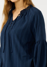 [Color: Midnight] A detail image of a blonde model wearing a blue long sleeve peasant blouse with a subtle checkered pattern. Featuring long bishop sleeves, ruffled neckline, tie detail, and button front. 