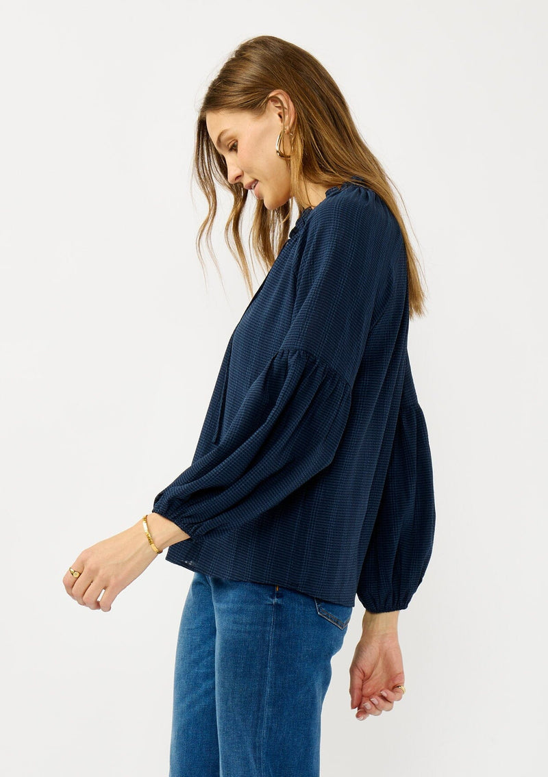 [Color: Midnight] A side facing image of a blonde model wearing a blue long sleeve peasant blouse with a subtle checkered pattern. Featuring long bishop sleeves, ruffled neckline, tie detail, and button front. 